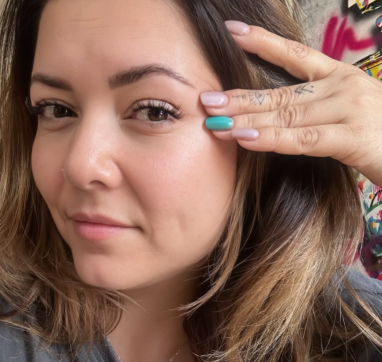 A woman with brown hair looks at the camera while gently pulling the skin near her eye with her fingers, showcasing her turquoise nail and hand tattoos. Her lashes appear fuller, hinting she is wearing Light Volume lash extensions from the Lash Link by HERA. The background is slightly blurred.